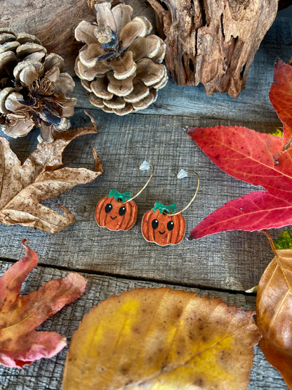A pair of smiley pumpkin mini hoop earrings arranged on rustic wood among red and yellow leaves, pine cones and bark.