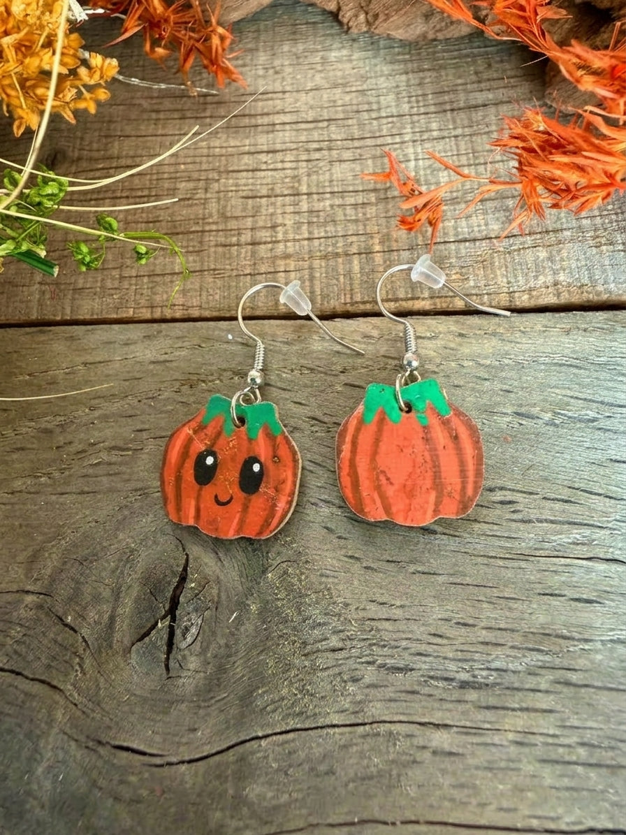 Silver hook pumpkin cork earrings showing the smiling face and painted reverse, arranged on wooden boards beneath autumn foliage