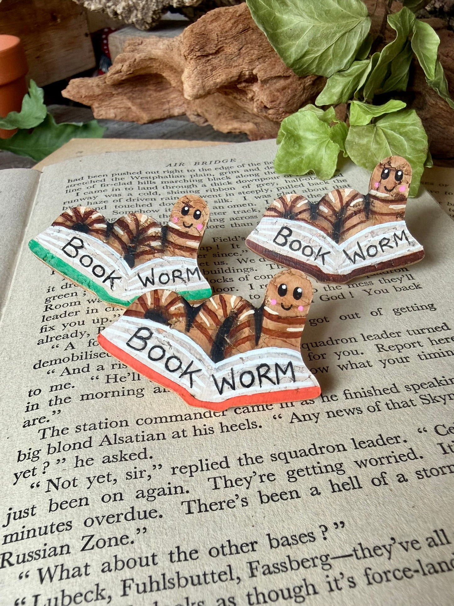 Natural bookworm cork pin badges in green, orange and brown book colourways on a vintage open book with driftwood and ivy — handpainted sustainable cork badges handmade in Suffolk UK