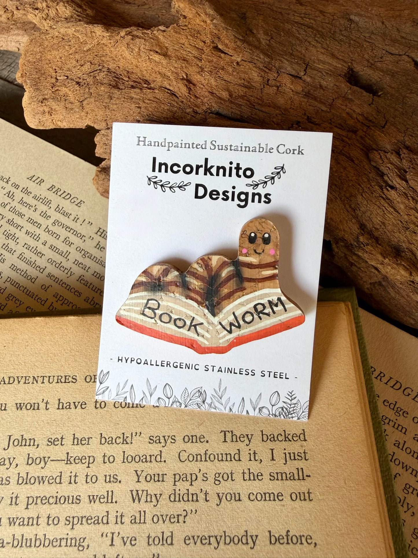 Natural bookworm cork pin badge on Incorknito Designs branded card against driftwood and open book — handpainted sustainable cork badge