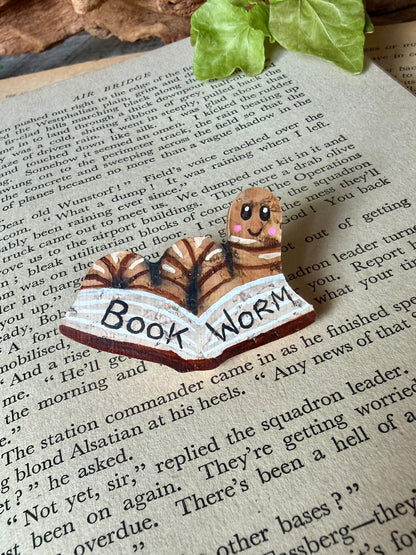 Natural bookworm cork pin badge with a cute smiley face with brown book edging on a vintage open book with driftwood and ivy backdrop — handpainted worm reading a book