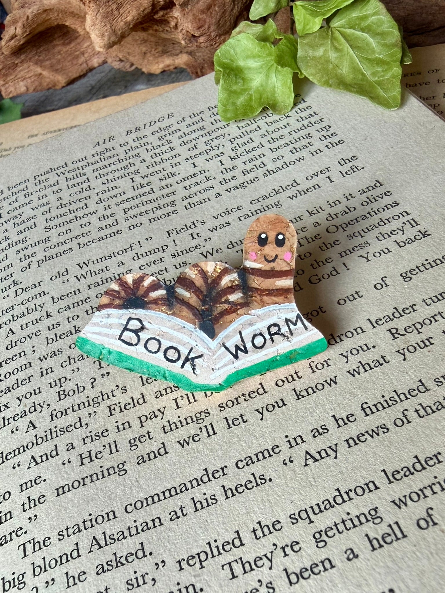Natural bookworm cork pin badge with green book on a vintage open book with driftwood and ivy backdrop — handpainted worm reading a book