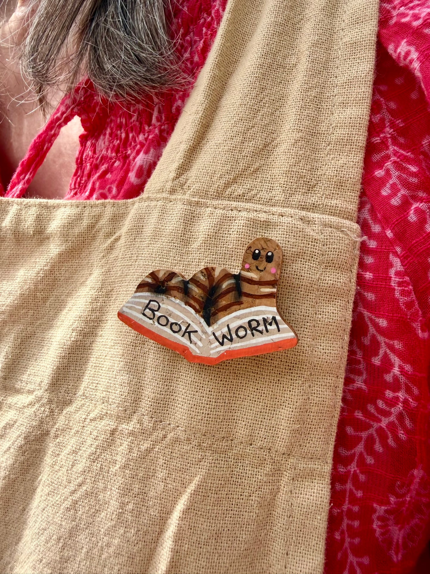 Natural bookworm cork pin badge pinned to a linen apron pocket — handpainted worm reading an open book, sustainable cork badge