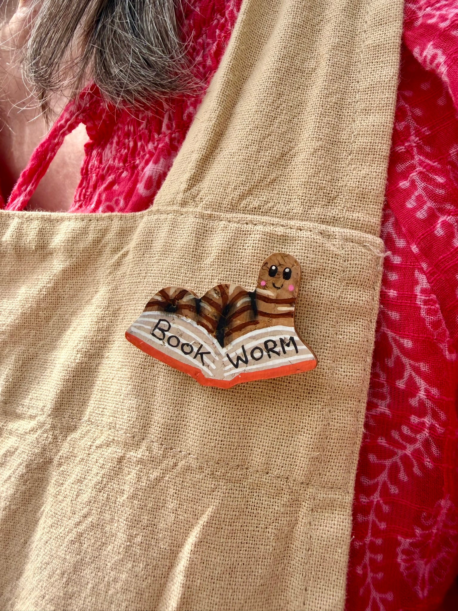 Natural bookworm cork pin badge pinned to a linen apron pocket — handpainted worm reading an open book, sustainable cork badge