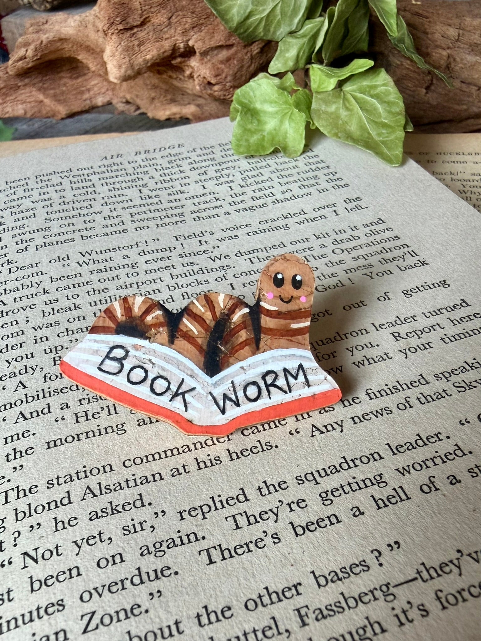 Natural bookworm cork pin badge with orange book on a vintage open book with driftwood and ivy backdrop — handpainted worm reading a book