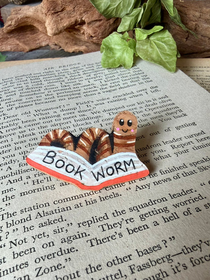 Natural bookworm cork pin badge with orange book on a vintage open book with driftwood and ivy backdrop — handpainted worm reading a book