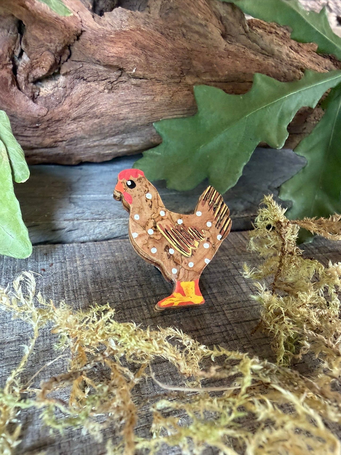 Natural chicken cork pin badge angled on weathered wood with driftwood, dried moss and oak leaves — handpainted golden-brown hen with white polka dots