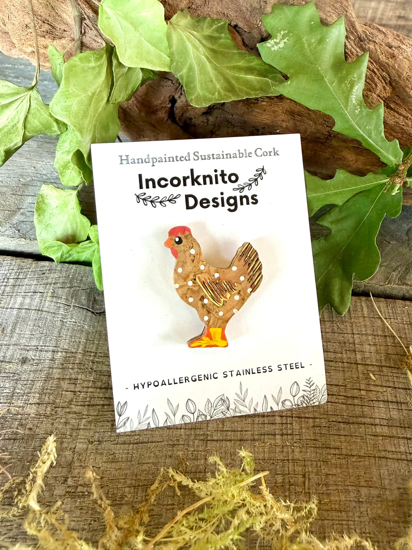 Natural chicken cork pin badge on Incorknito Designs branded card against driftwood and oak leaves — handpainted golden-brown hen with white polka dots, handmade in Suffolk UK