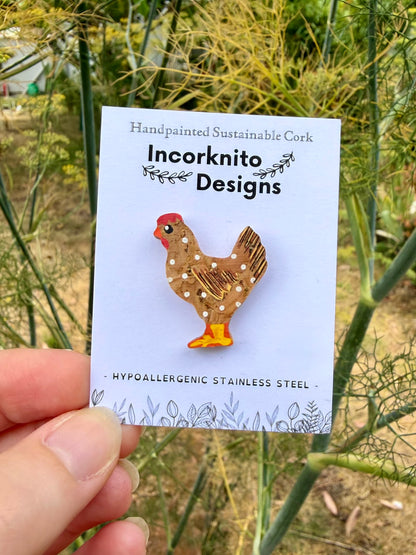 Natural chicken cork pin badge on Incorknito Designs branded card held against garden fennel — handpainted golden-brown hen with white polka dots, sustainable cork badge