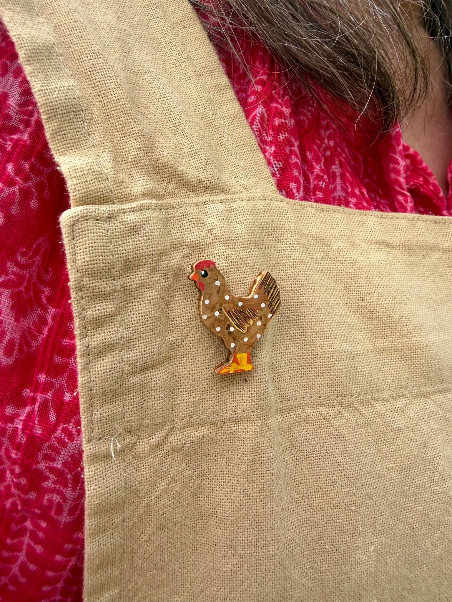 Natural chicken cork pin badge pinned to a natural linen apron pocket — handpainted golden-brown hen with white polka dots and gold tail feathers, sustainable cork badge handmade in Suffolk UK