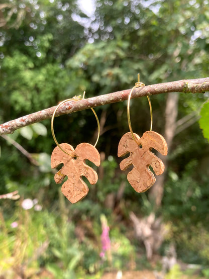 Natural cork monstera earrings on gold hoops suspended from a lichen-covered branch — handmade sustainable plant jewellery with garden foxgloves in the background