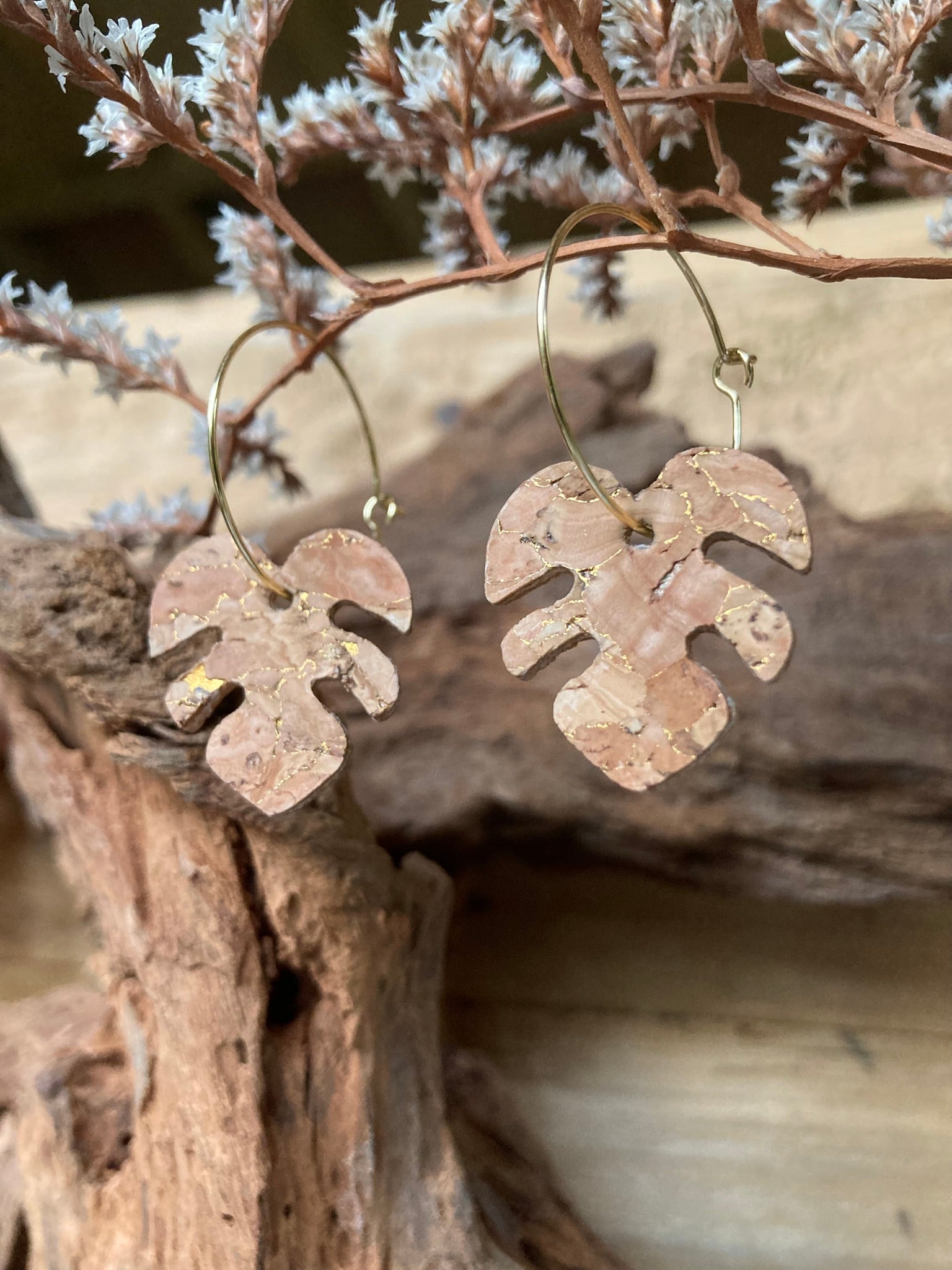 Gold hoop natural cork monstera earrings resting against driftwood with dried white botanical sprigs — eco-friendly plant-inspired jewellery