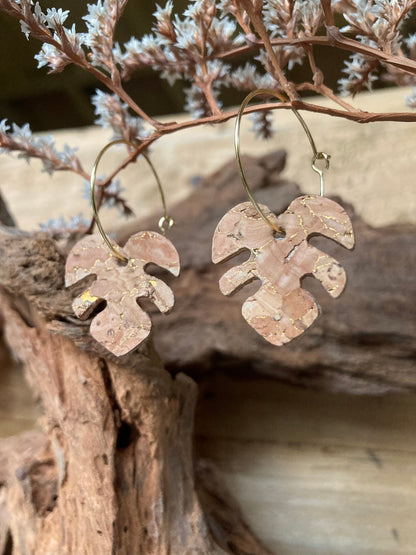 Gold hoop natural cork monstera earrings resting against driftwood with dried white botanical sprigs — eco-friendly plant-inspired jewellery