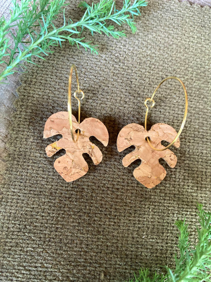 Flat lay of natural cork monstera earrings on gold hoops on olive textured fabric with sprigs of green cypress — handmade plant jewellery Suffolk UK