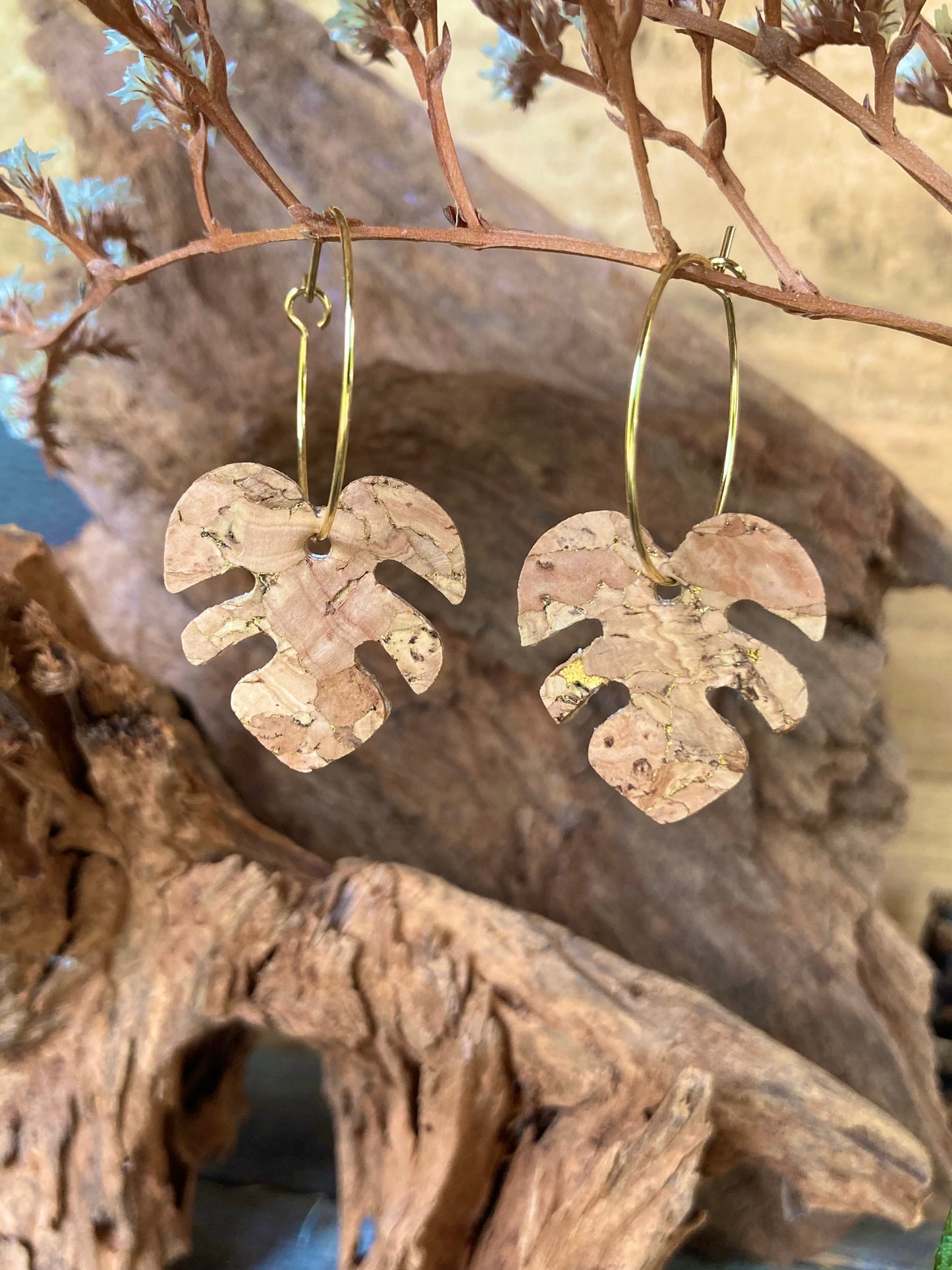 Natural cork monstera earrings on gold hoops hanging from a russet branch against weathered driftwood — gold fleck cork texture catching warm light