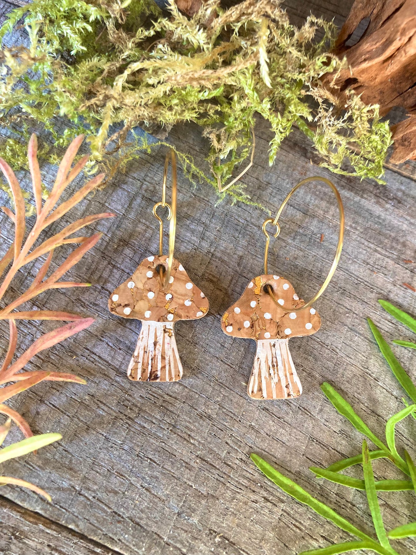 Natural toadstool cork large hoop earrings flat lay on weathered wood with moss and red botanicals — gold hoops with natural cork cap and white spot detail, handmade in Suffolk UK