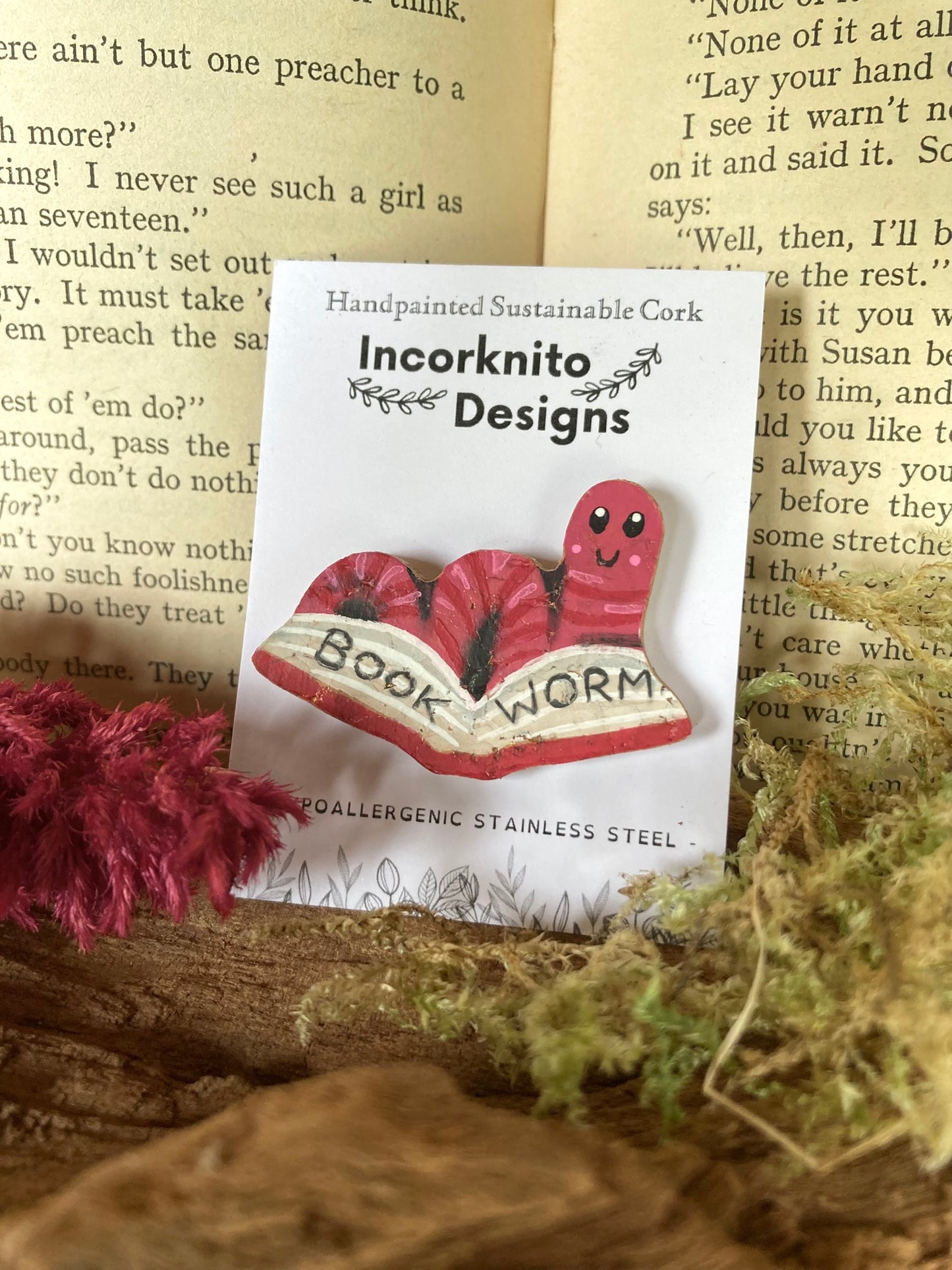 Pink bookworm cork pin badge on Incorknito Designs branded card against a moss and open book background — handpainted sustainable badge handmade in Suffolk UK