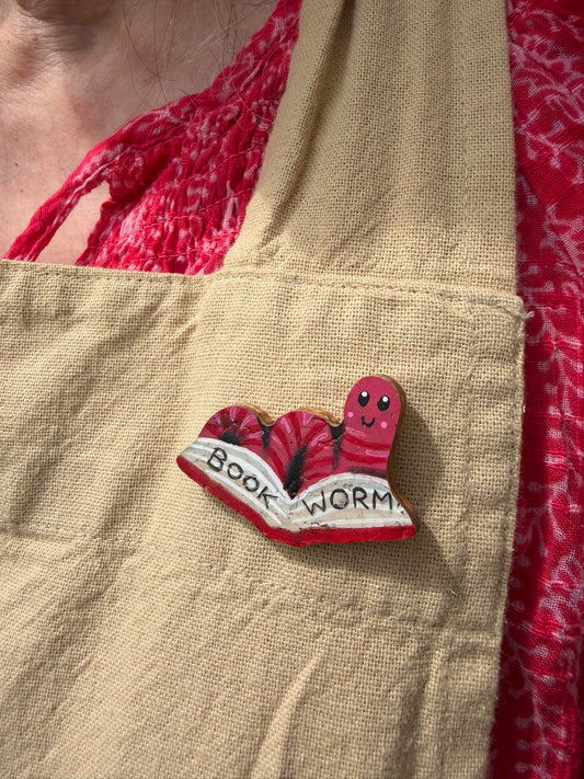 Pink bookworm cork pin badge pinned to a linen apron — handpainted worm reading an open book, sustainable cork badge handmade in Suffolk UK