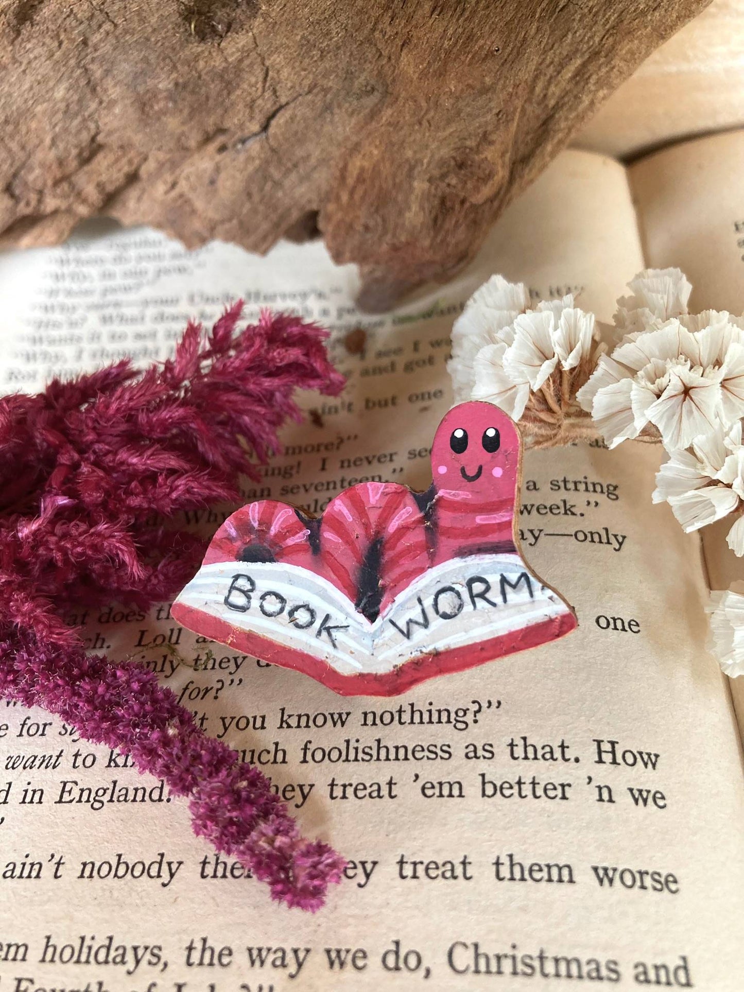Handpainted pink bookworm cork pin badge resting on an open book with dried pink and white botanicals and driftwood — eco-friendly badge handmade in Suffolk UK