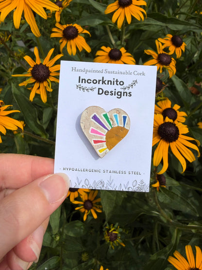 Rainbow sun heart cork pin badge on Incorknito Designs branded card held against yellow rudbeckia flowers — handpainted multicolour sunrise heart, sustainable cork badge handmade in Suffolk UK
