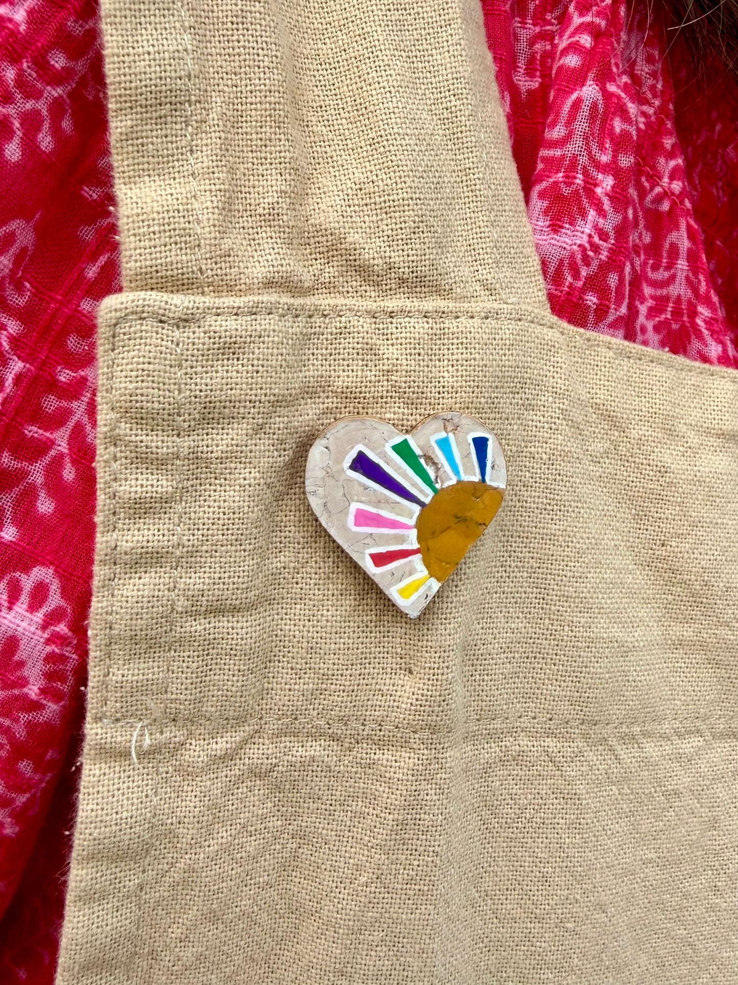 Rainbow sun heart cork pin badge pinned to a linen apron — handpainted multicolour sunrise heart with golden sun, sustainable cork badge