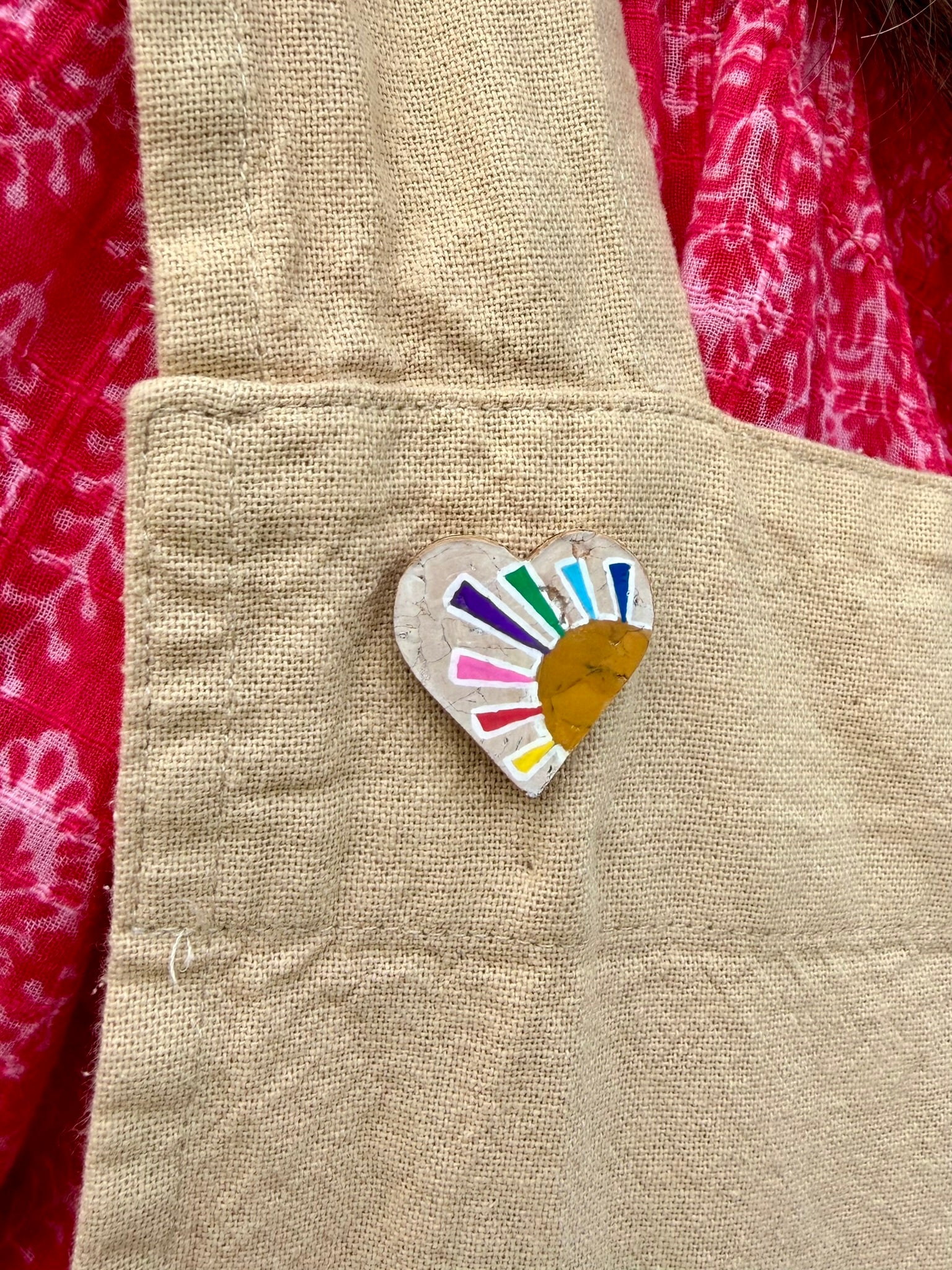 Rainbow sun heart cork pin badge pinned to a linen apron — handpainted multicolour sunrise heart with golden sun, sustainable cork badge