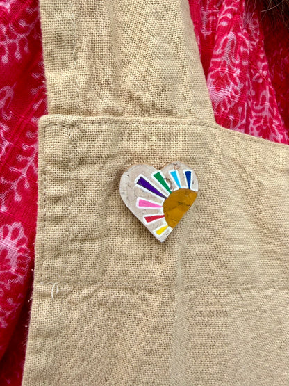 Rainbow sun heart cork pin badge pinned to a linen apron — handpainted multicolour sunrise heart with golden sun, sustainable cork badge