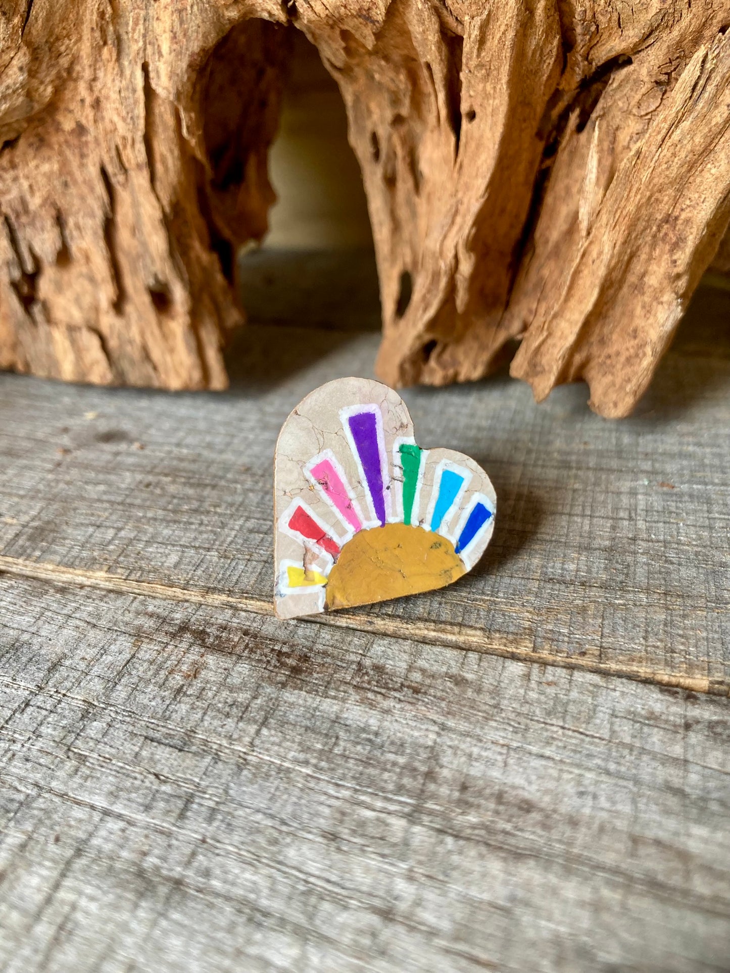 Heart-shaped rainbow sun cork pin badge propped against driftwood at an angle — handpainted multicoloured rays, eco-friendly badge handmade in UK