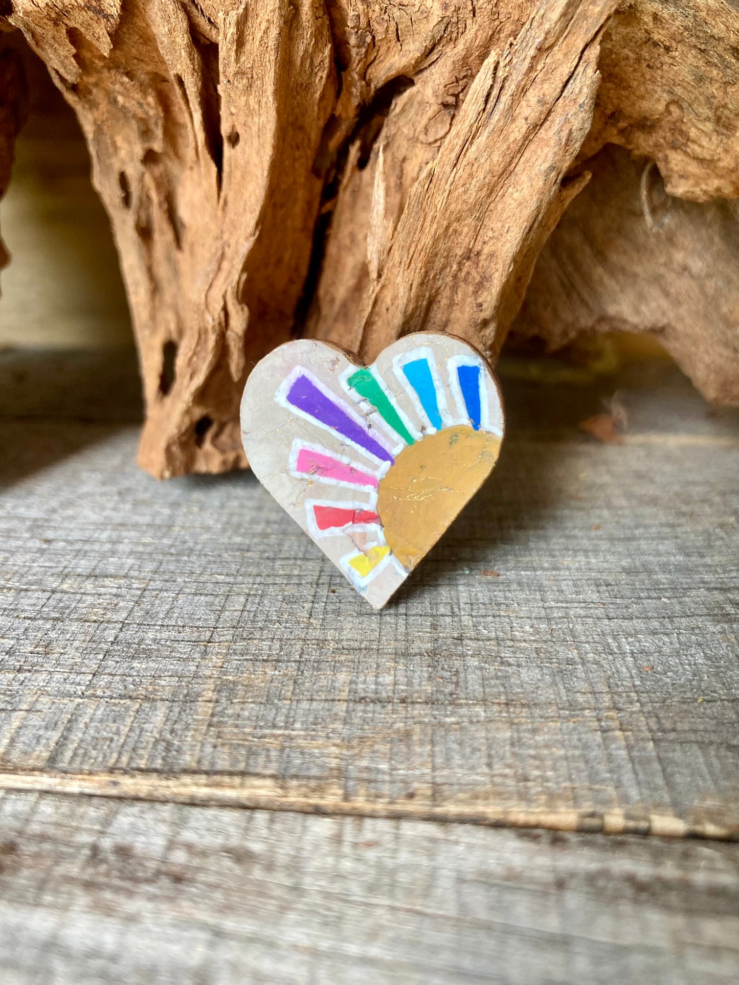 Handpainted rainbow sun cork pin badge propped against driftwood — heart-shaped badge with multicoloured rays and golden sun, handmade in Suffolk UK