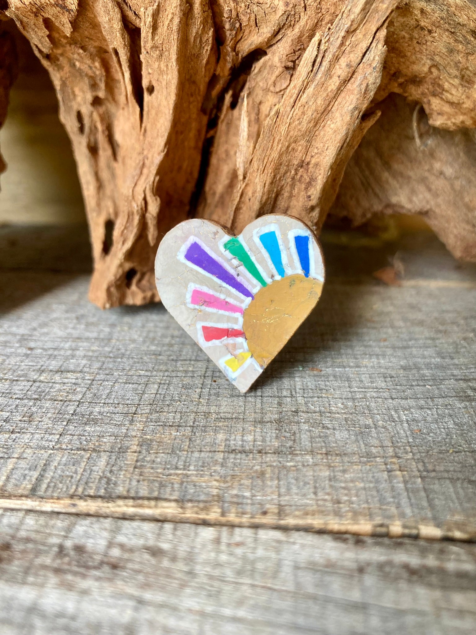 Handpainted rainbow sun cork pin badge propped against driftwood — heart-shaped badge with multicoloured rays and golden sun, handmade in Suffolk UK