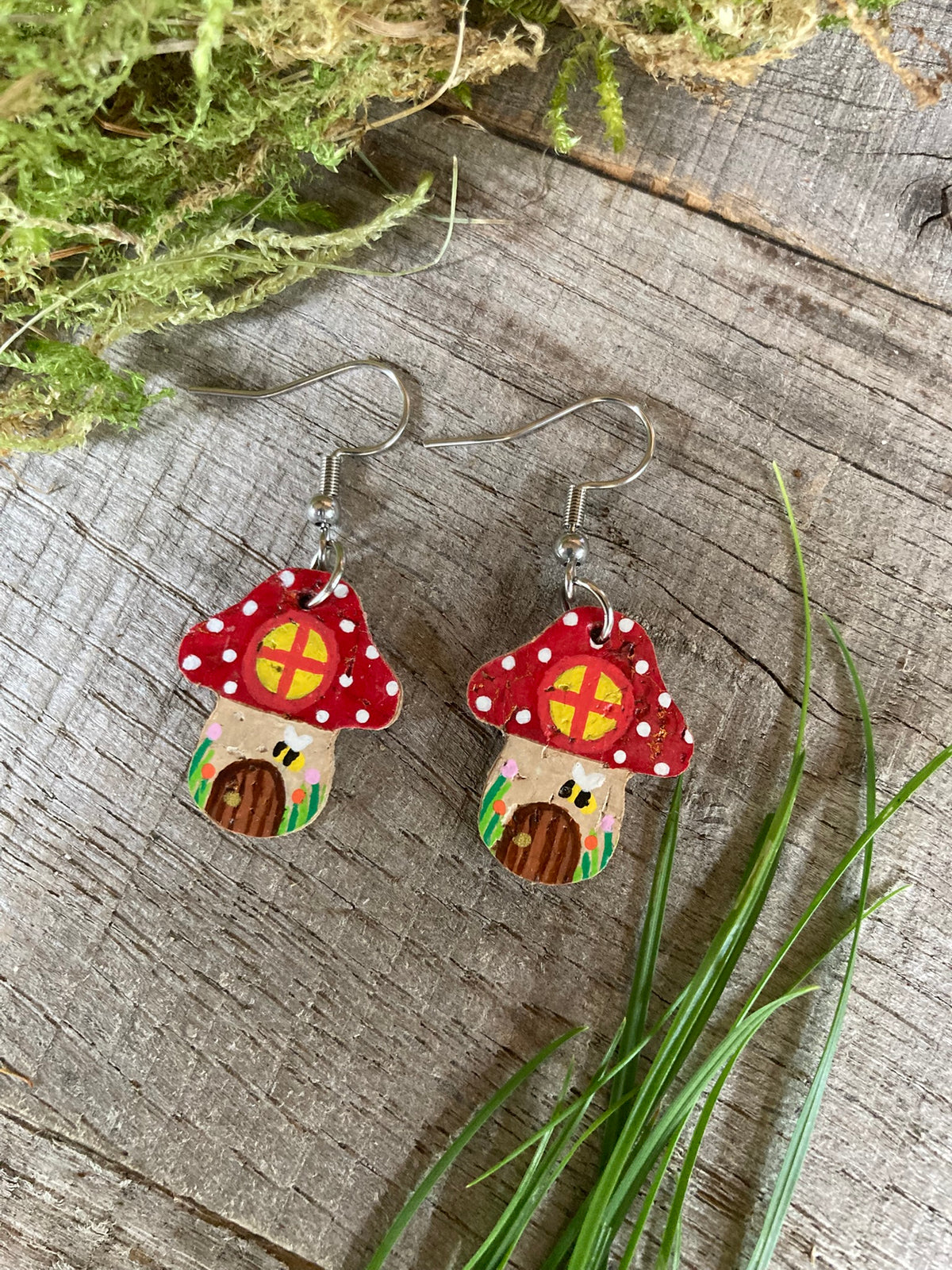 Red Fairy House Toadstool Earrings on hooks, displayed on a wooden surface with natural greenery