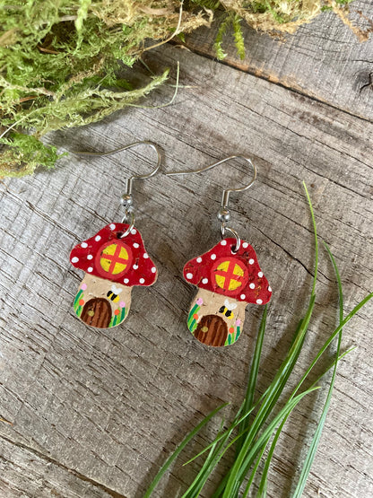 Red Fairy House Toadstool Earrings on hooks, displayed on a wooden surface with natural greenery