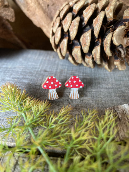Red toadstool cork stud earrings with white polka dots on a wooden surface with pine cone and moss