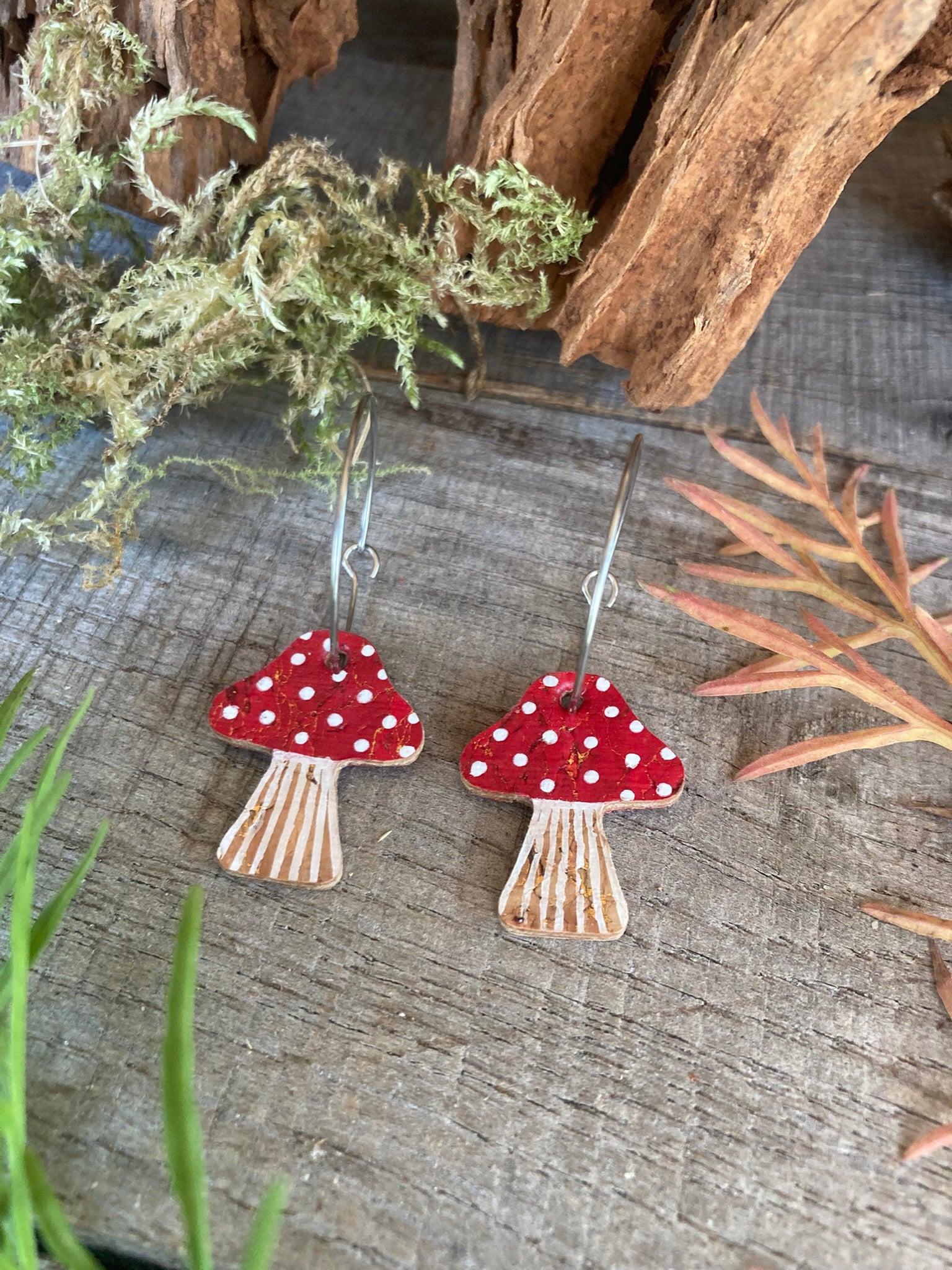Red toadstool cork large hoop earrings flat lay on weathered wood with greenery — handpainted fly agaric cap with white spot detail