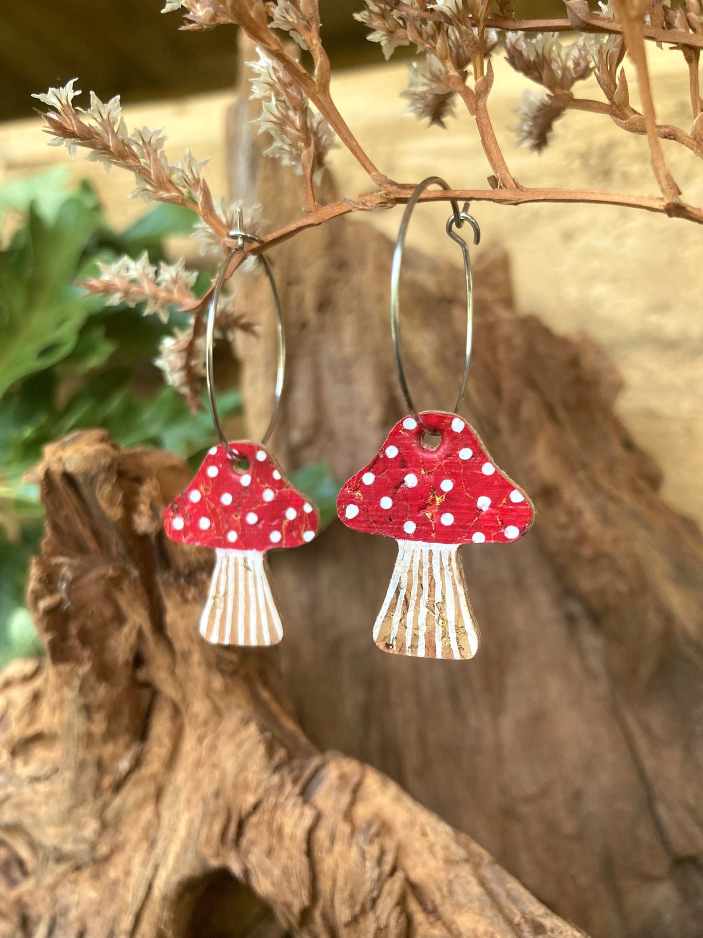Red toadstool cork large hoop earrings hanging from driftwood with botanicals — showing the hoop drop and handpainted fly agaric design, handmade in Suffolk UK