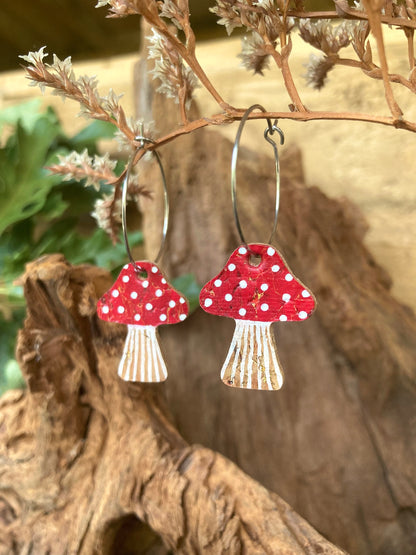 Red toadstool cork large hoop earrings hanging from driftwood with botanicals — showing the hoop drop and handpainted fly agaric design, handmade in Suffolk UK