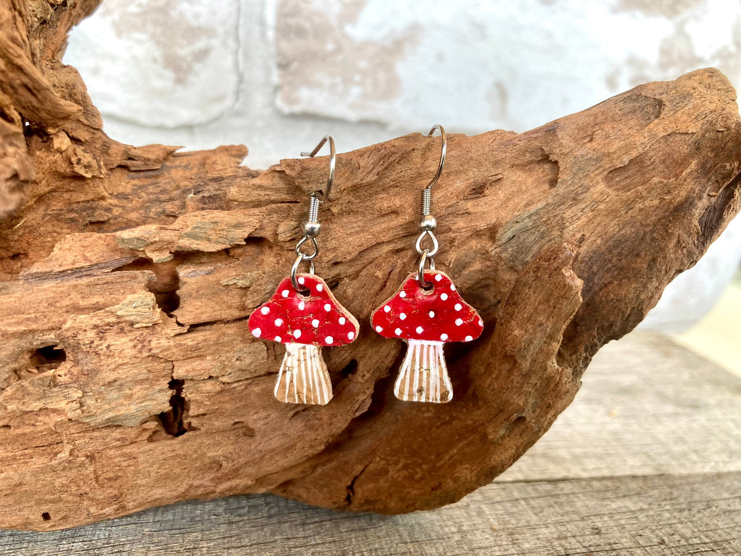 Red toadstool cork medium earrings hanging from driftwood — silver hooks showing the handpainted fly agaric cap and striped stem