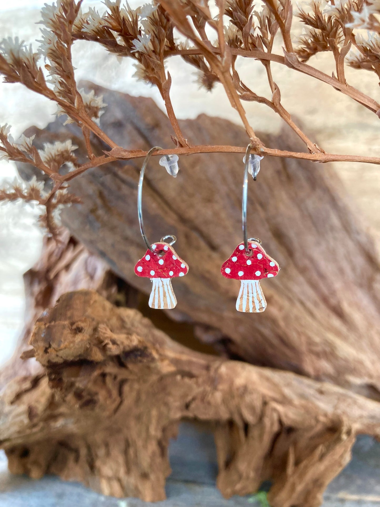 Red toadstool cork mini hoop earrings hanging from dried russet botanicals — handpainted fly agaric design with white spots on a rich red cap