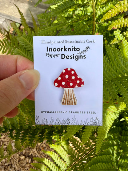 Red toadstool cork pin badge on Incorknito Designs branded card held against green ferns — handpainted fly agaric, sustainable cork badge handmade in Suffolk UK