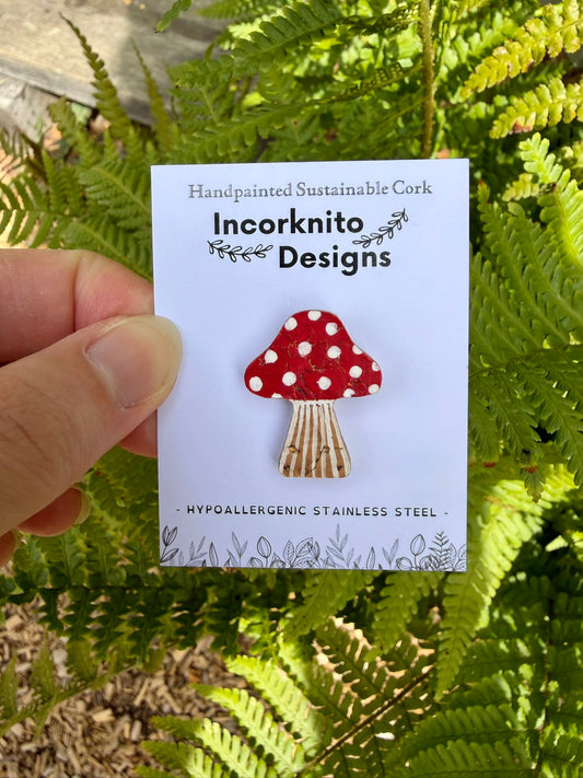 Red toadstool cork pin badge on Incorknito Designs branded card held against green ferns — handpainted fly agaric, sustainable cork badge handmade in Suffolk UK