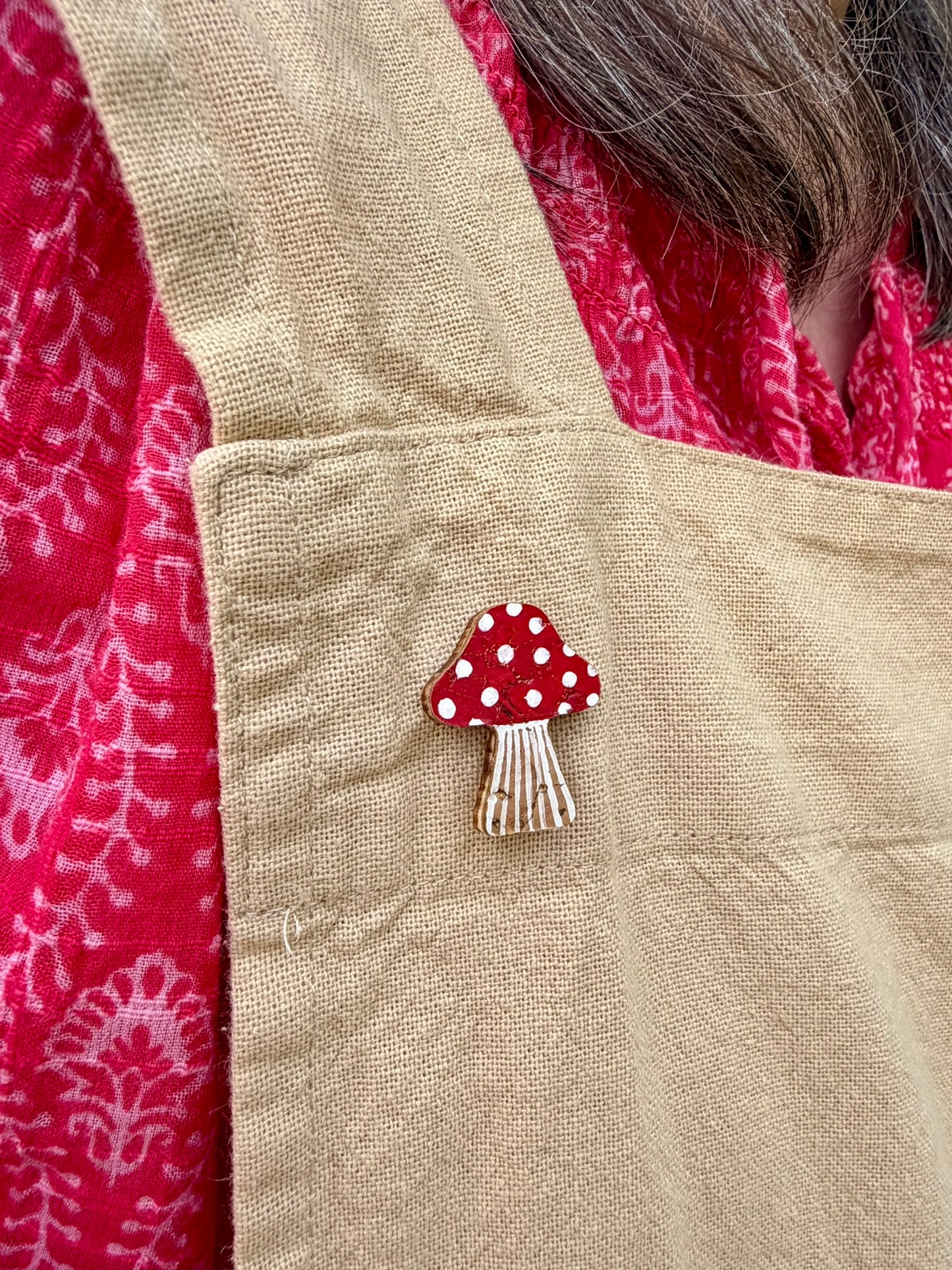 Red toadstool cork pin badge pinned to a linen apron pocket — handpainted fly agaric with white spots, sustainable cork badge