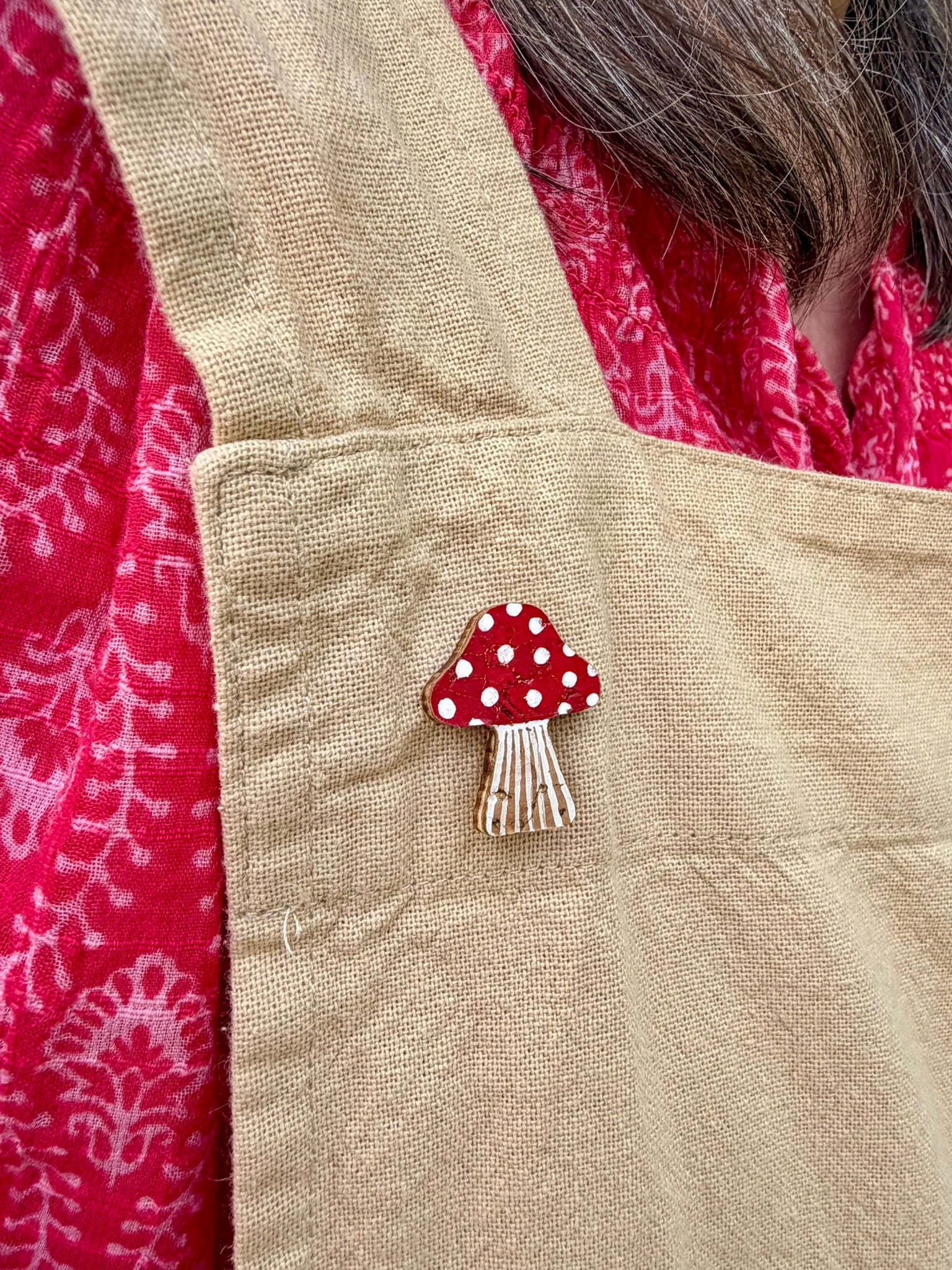 Red toadstool cork pin badge pinned to a linen apron pocket — handpainted fly agaric with white spots, sustainable cork badge