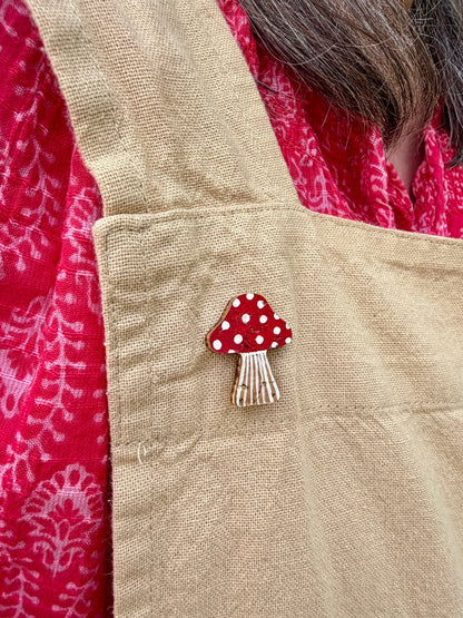 Red toadstool cork pin badge pinned to a linen apron pocket — handpainted fly agaric with white spots, sustainable cork badge