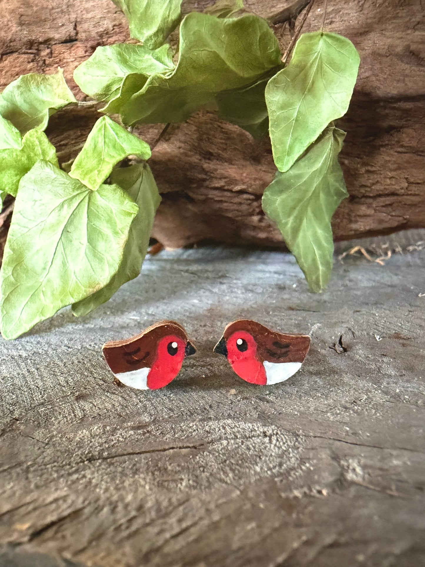 Handpainted robin cork stud earrings with vivid red breast and brown wings on a driftwood surface with ivy