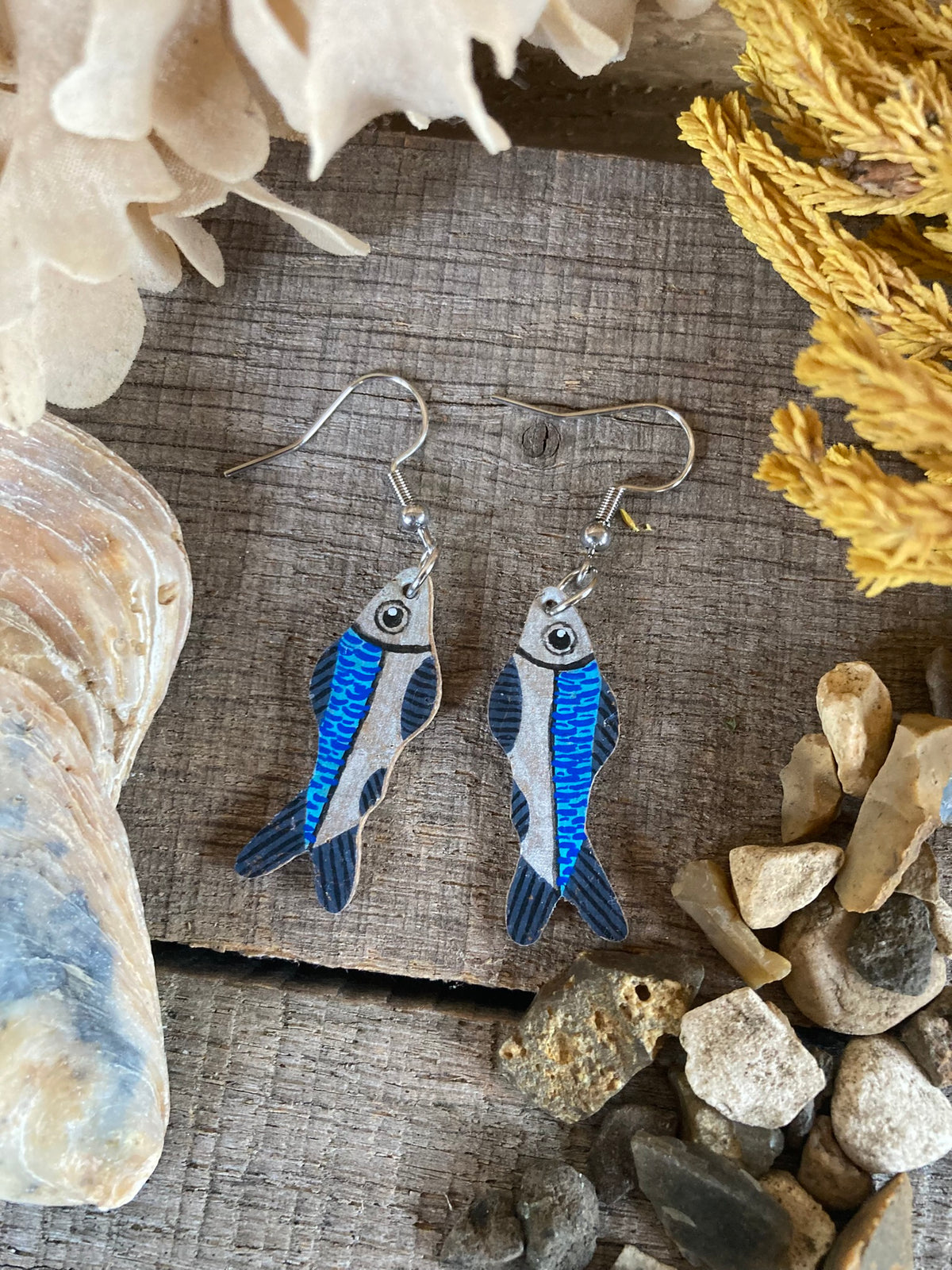 Handpainted sardine cork earrings on silver hooks, flat lay on driftwood with shells and coastal pebbles