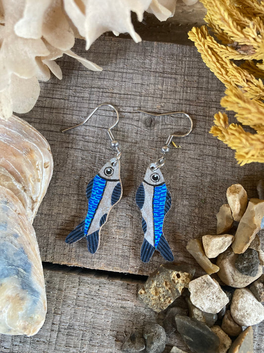 Handpainted sardine cork earrings on silver hooks, flat lay on driftwood with shells and coastal pebbles