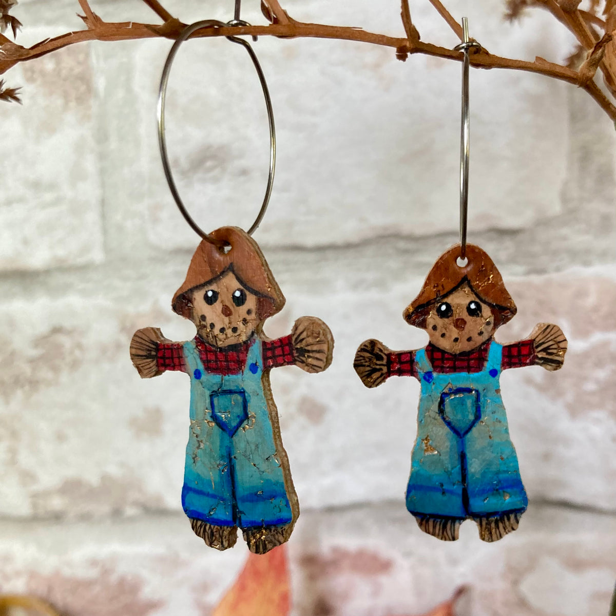 Handpainted scarecrow cork earrings on large silver hoops, hanging against a white brick wall with autumn foliage