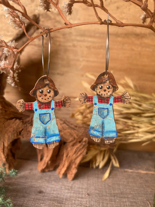 Handpainted cork scarecrow earrings on silver hoops, hanging from a dried branch against a warm autumnal background. Red tartan shirt, blue denim dungarees and a brown floppy hat