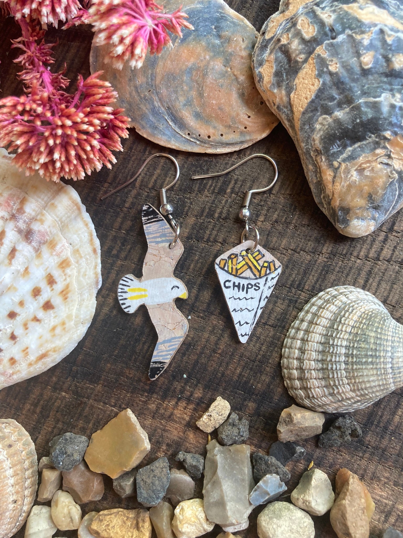 Handpainted seagull and chips cork earrings on silver hooks flat lay on weathered wood with seashells, stones and dried red botanicals — eco-friendly coastal jewellery