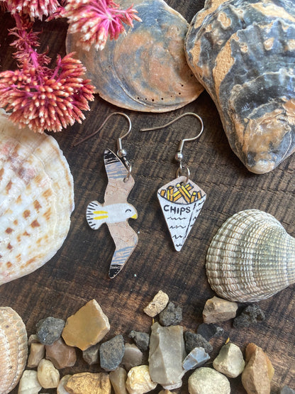 Handpainted seagull and chips cork earrings on silver hooks flat lay on weathered wood with seashells, stones and dried red botanicals — eco-friendly coastal jewellery